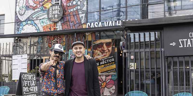Distrito San Felipe fachada de Casa Valhalla pintada con grafiti y rejas de color negro. Hay dos personas sonriendo a la cámara delante de un aviso comercial con la figura de Hector Lavoe y otro de cerveza.