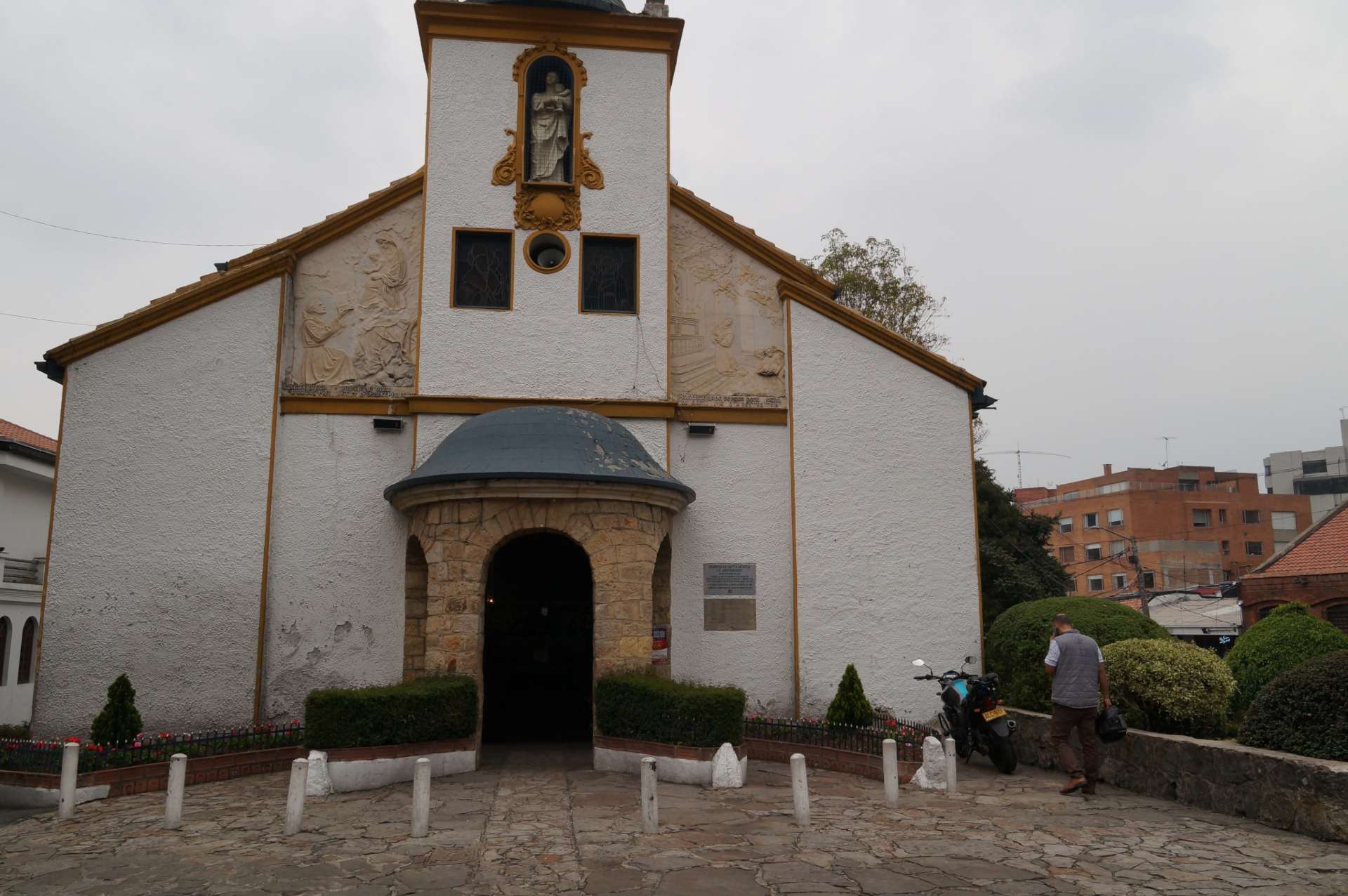 Iglesia de Santa Mónica, fachada de día con cielo gris. Paredes blancas con dos ventanas y entrada de piedra en forma circular como un quiosco con cúpula gris y dos pequeños pinos a los costados. Al lado izquierdo hay un hombre y una moto estacionada.
