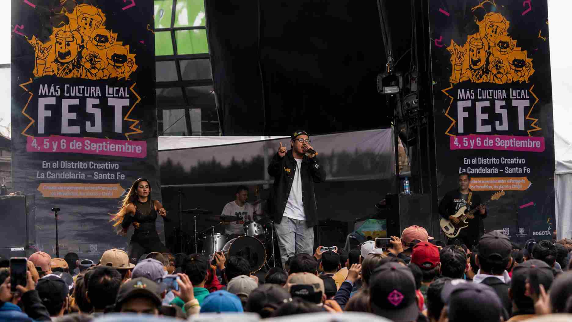 Cantante en escenario durante el Más Cultura Local Fest, ante una multitud de asistentes con banderas publicitarias. (119 caracteres)
