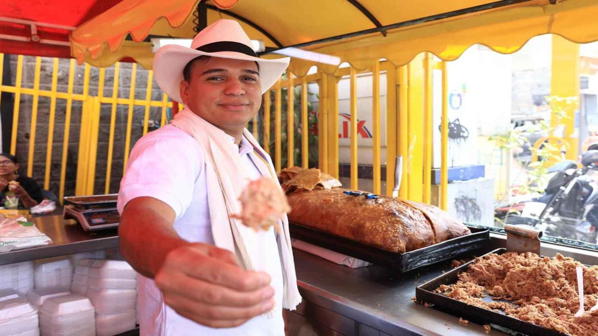 Un hombre con sombrero y raboegallo ofrece una porción de lechona en primer plano junto a su puesto de comida.