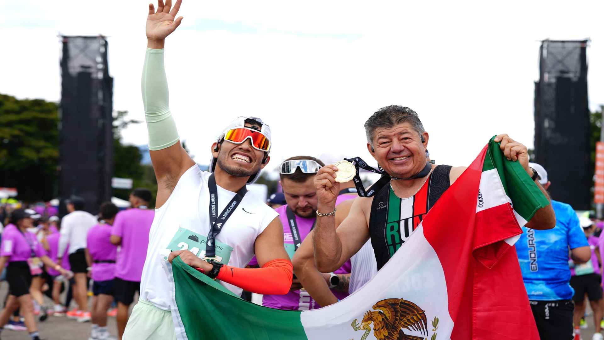 Dos corredores mexicanos celebran felices en la MMB 2025 sosteniendo su bandera nacional y mostrando su medalla.
