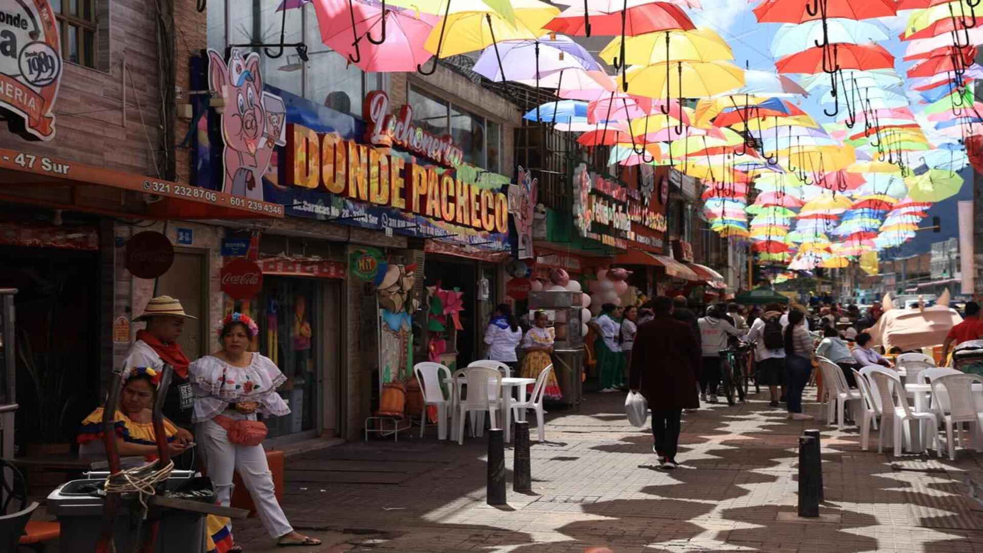 Calle peatonal decorada con sombrillas de colores en el techo y locales con letreros de lechonerías tradicionales.