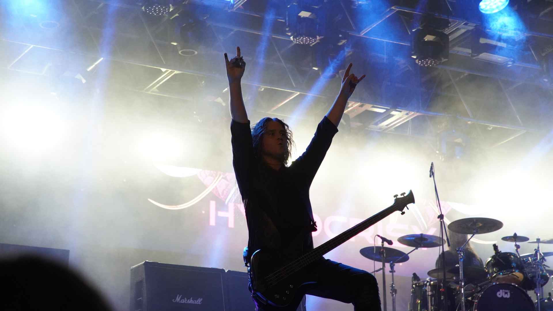 Músico en el escenario con los brazos en alto haciendo la señal del rock, luces de fondo y batería a la derecha.