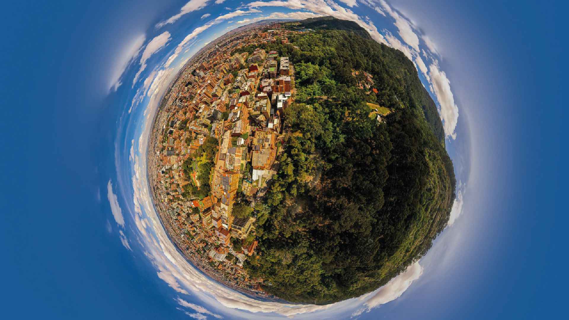 Fotografía panorámica en efecto ojo de pez de una ciudad rodeada por montañas verdes bajo un cielo azul.