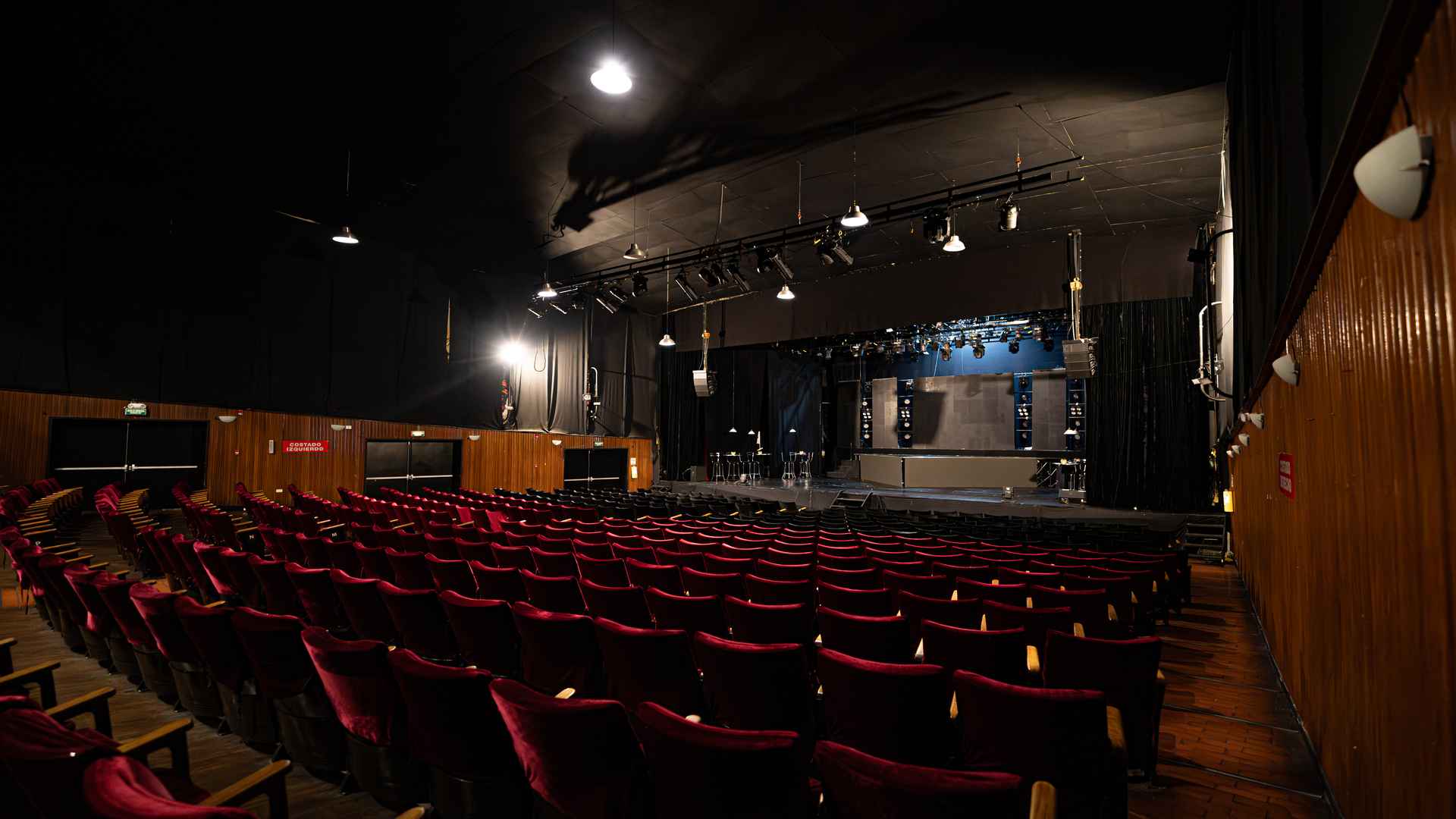 Vista amplia de la sala de teatro Arlequín con sillería de terciopelo rojo, paredes de madera y escenario iluminado.