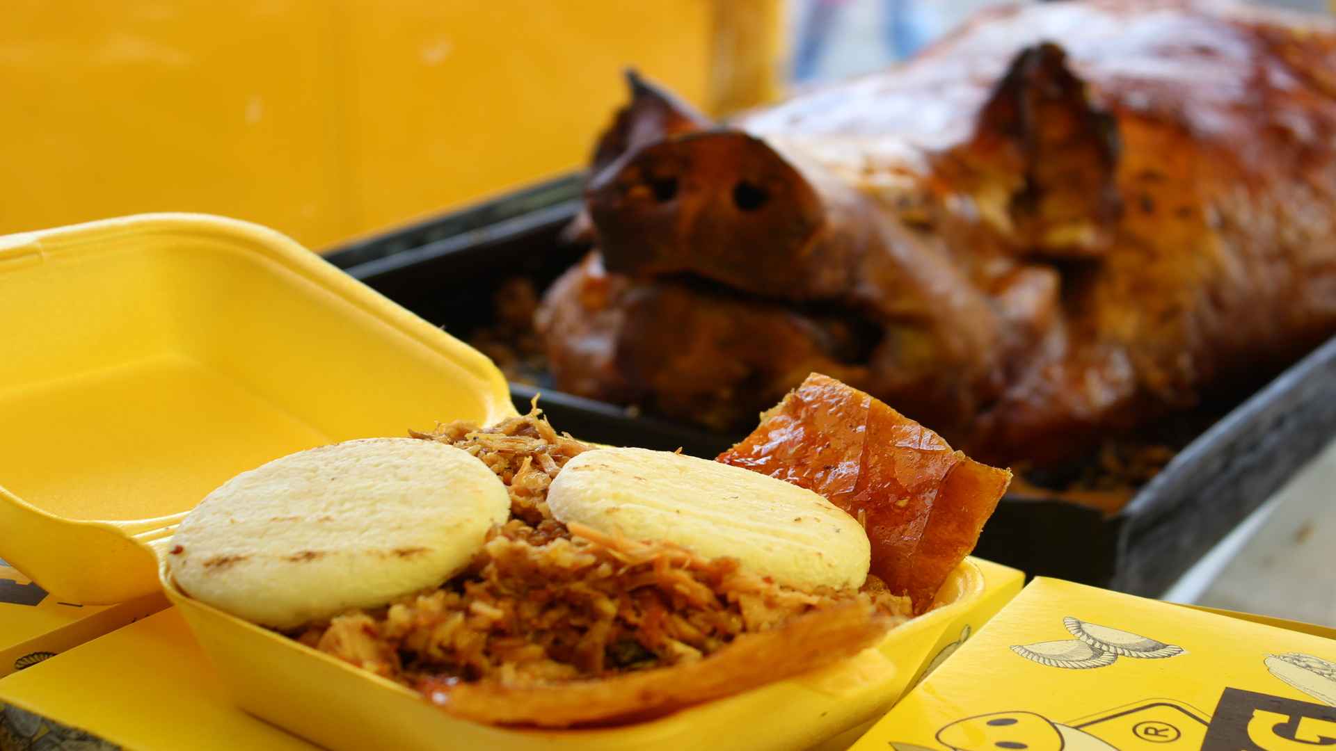 Un envase plástico amarillo con una porción de lechona y dos arepas frente a un cerdo horneado entero.