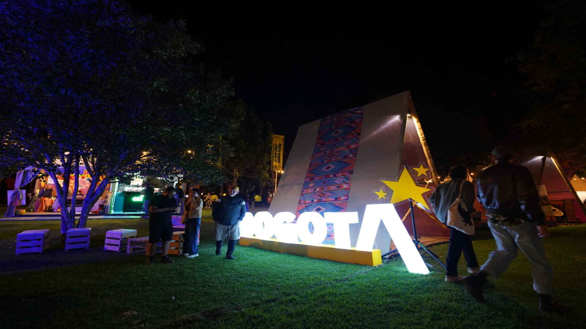 Letrero gigante e iluminado de Bogotá junto a una carpa decorada en un parque durante la noche con transeúntes.