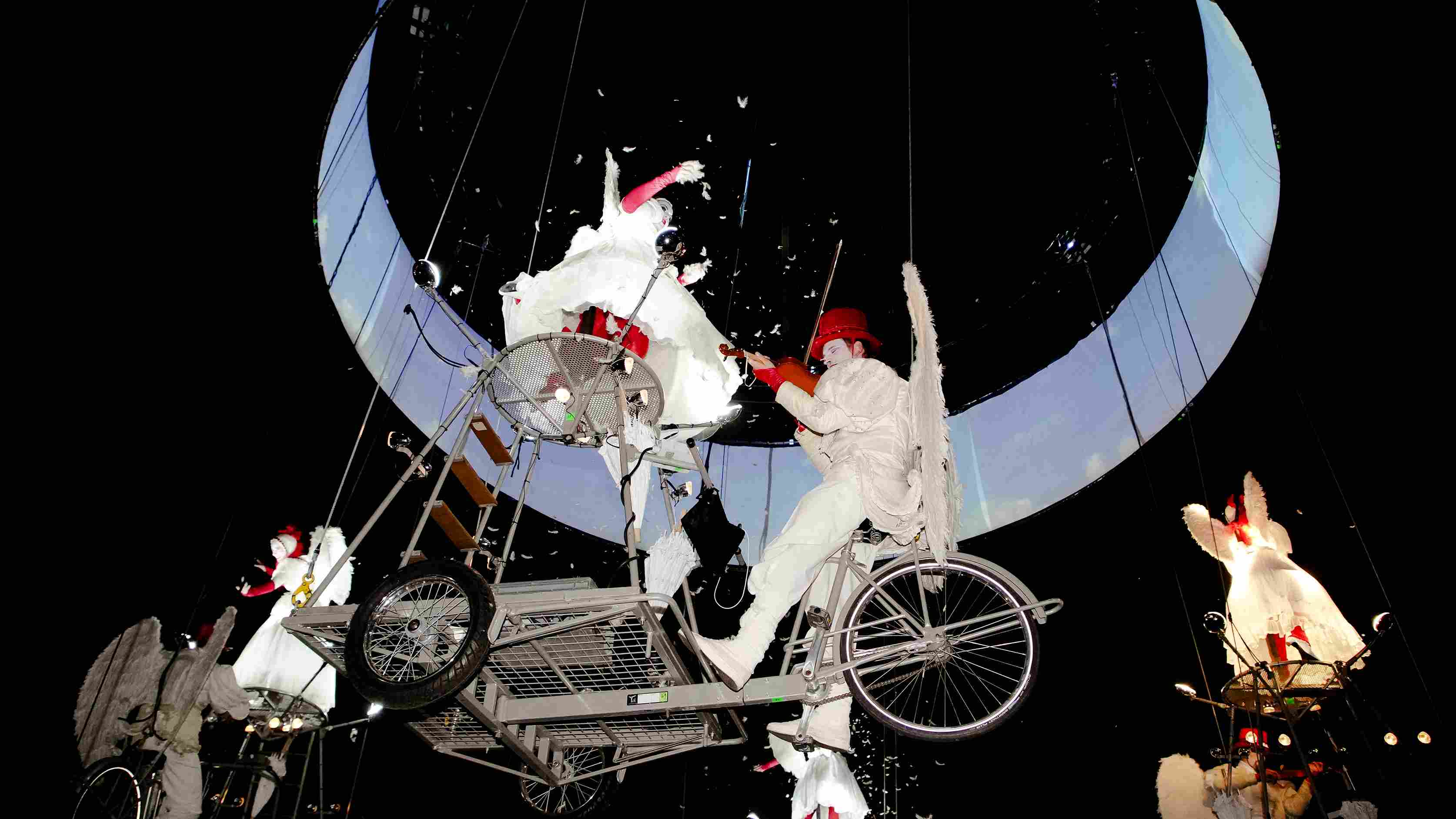 Estructura mecánica suspendida con músicos y figuras aladas en interacción, bajo una iluminación circular blanca. Fotografía Archivo IDT