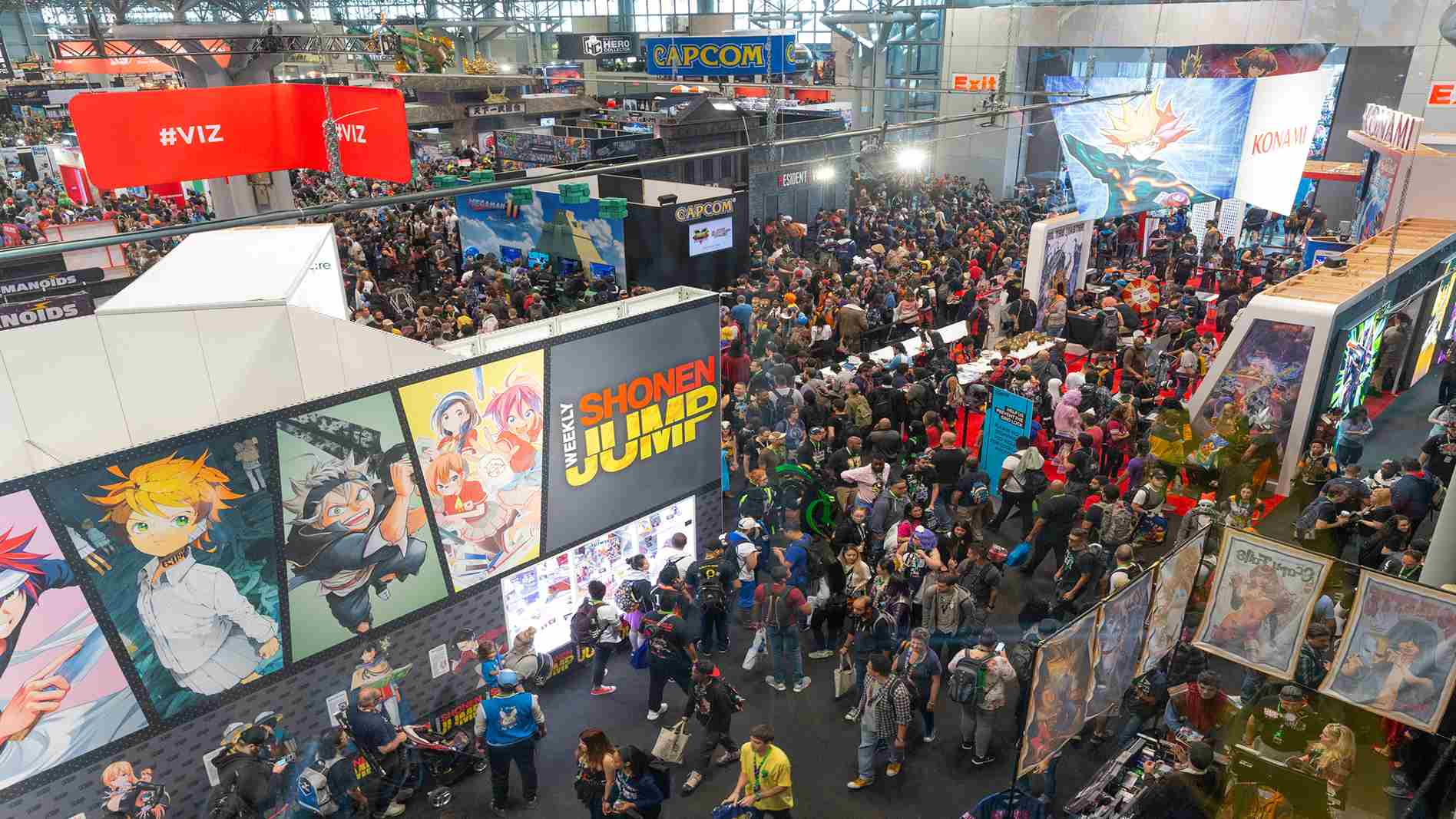 Vista aérea de un pabellón repleto de personas caminando entre estands con grandes pósteres de manga y videojuegos.