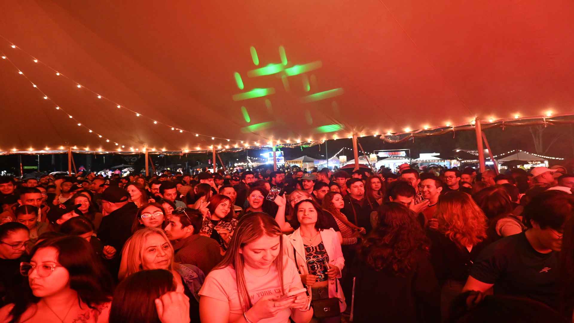 Gran multitud de personas reunidas bajo una carpa iluminada con luces cálidas y un reflector verde en el techo.