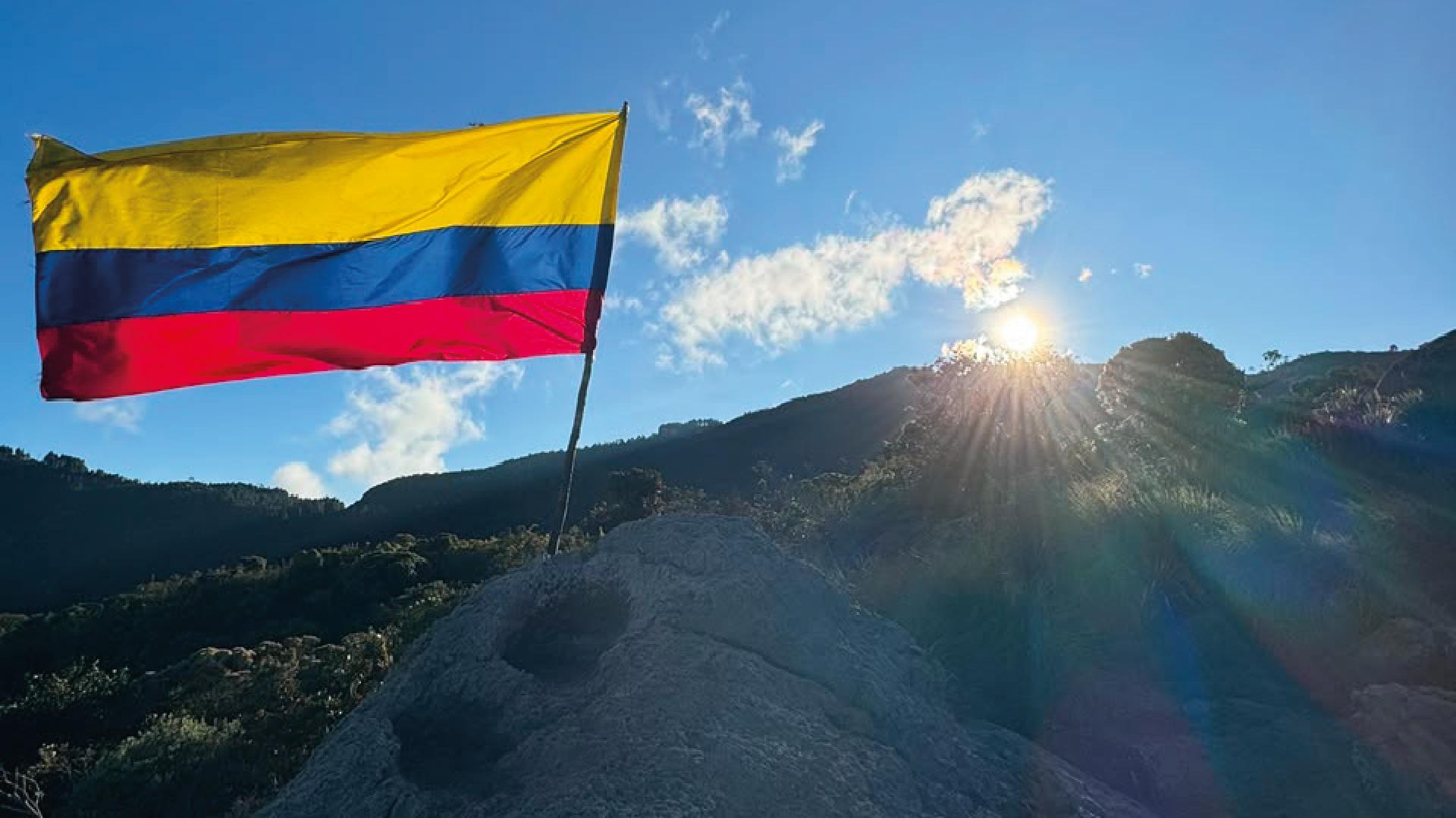 Bandera de Colombia ondeando sobre una gran roca en la cima de una montaña, con el sol resplandeciendo de fondo.