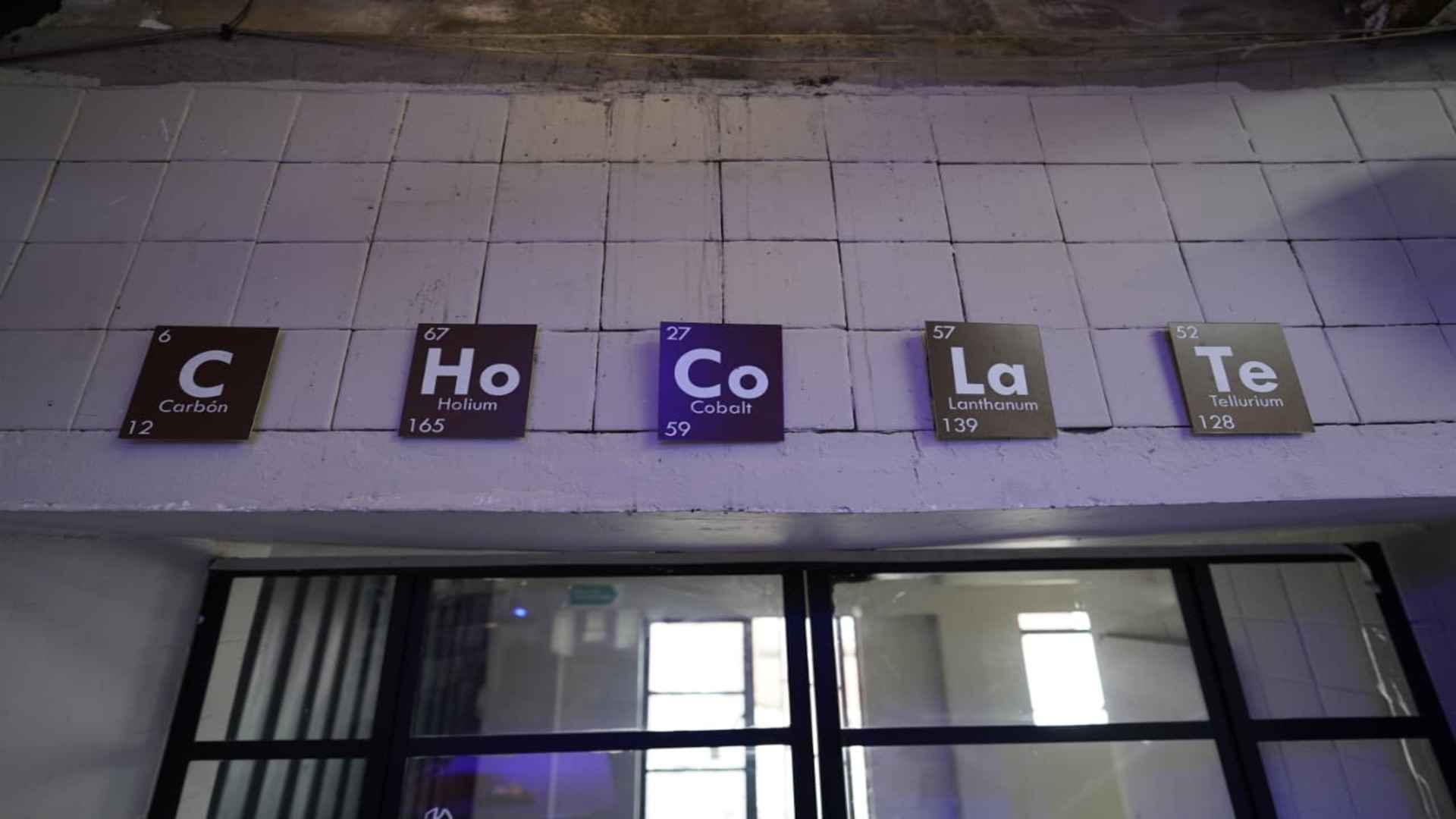 Letrero en pared de azulejos blancos que deletrea "Ch O Co La Te" simulando elementos de la tabla periódica de química.