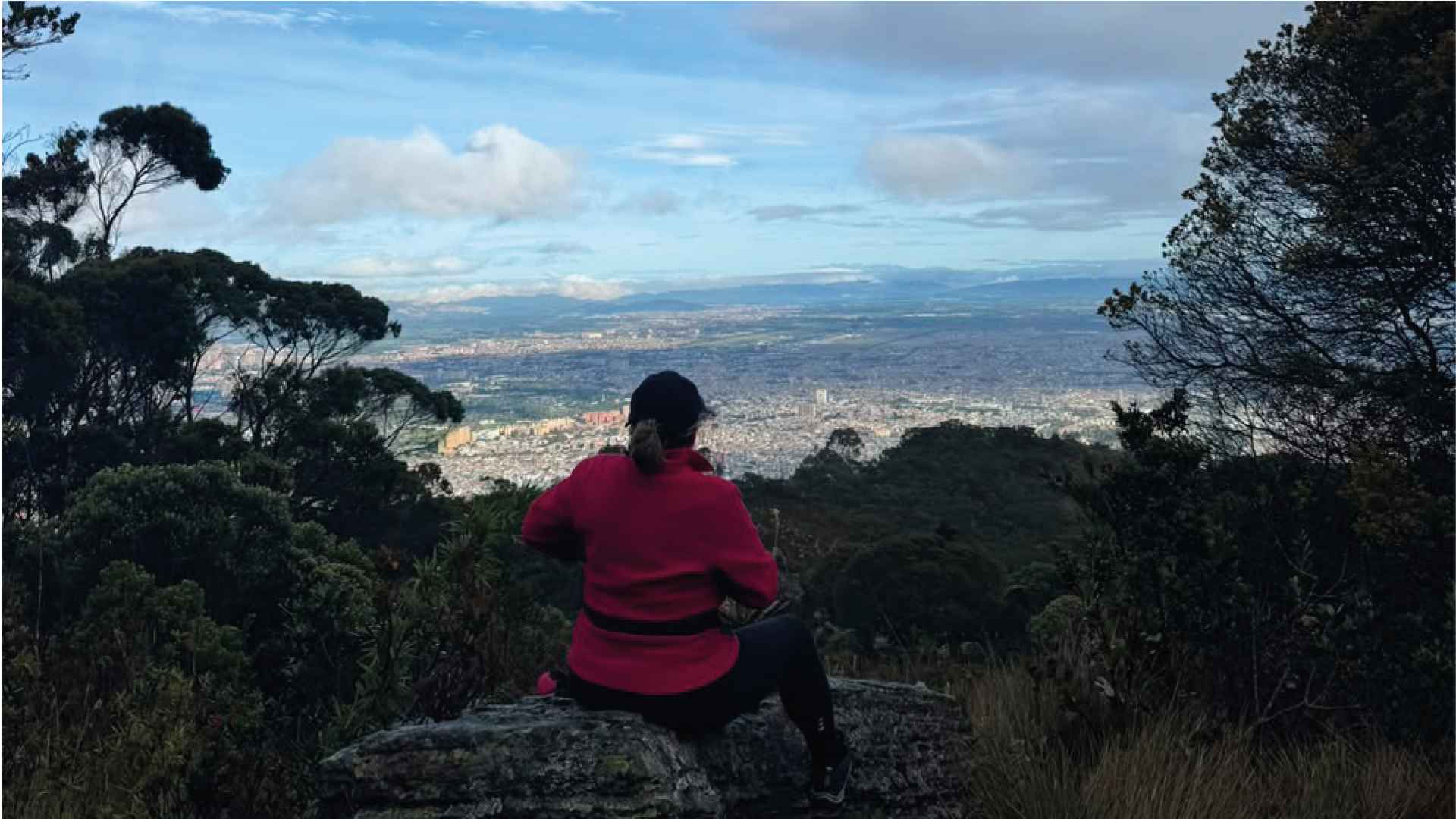Mujer de chaqueta roja sentada de espaldas sobre una roca, contemplando la vista panorámica de la ciudad.