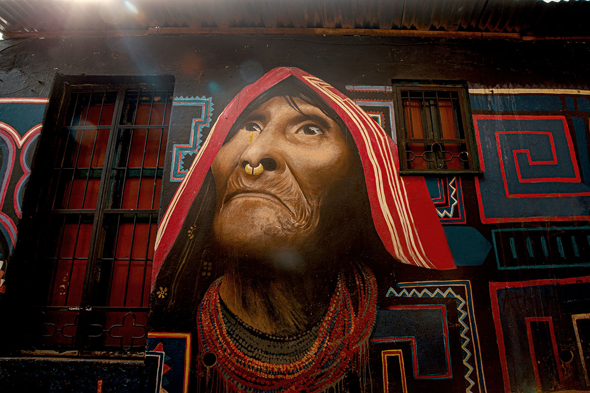 Grafiti de colores en fachada de casa con dibujo de rostro de indigena con collares. Dos ventanas en hierro pintadas de negro y rojo.