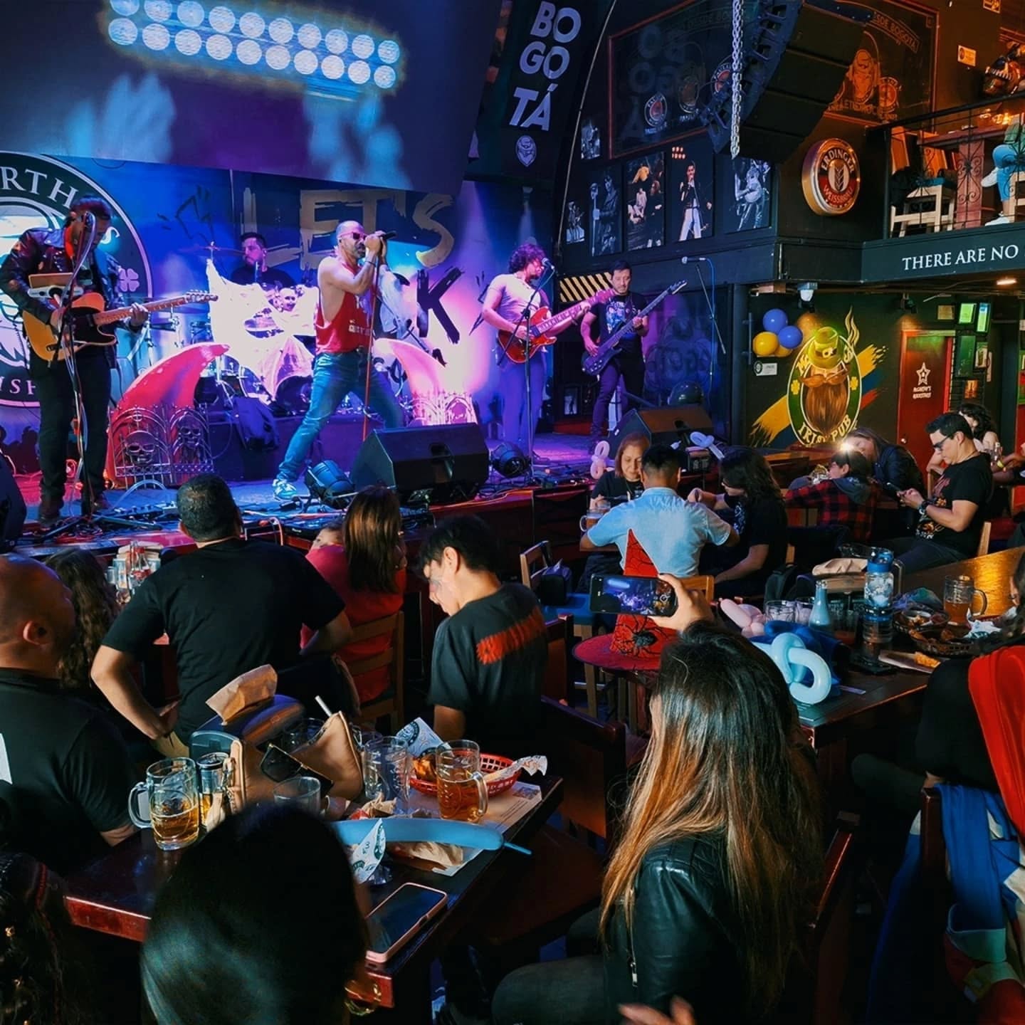 Bar de rock con banda de 5 integrantes en escenario. El lugar está lleno con público sentado bebiendo cerveza en mesas al frente de la tarima. El lugar está rodeado de fotografías de bandas rockeras.