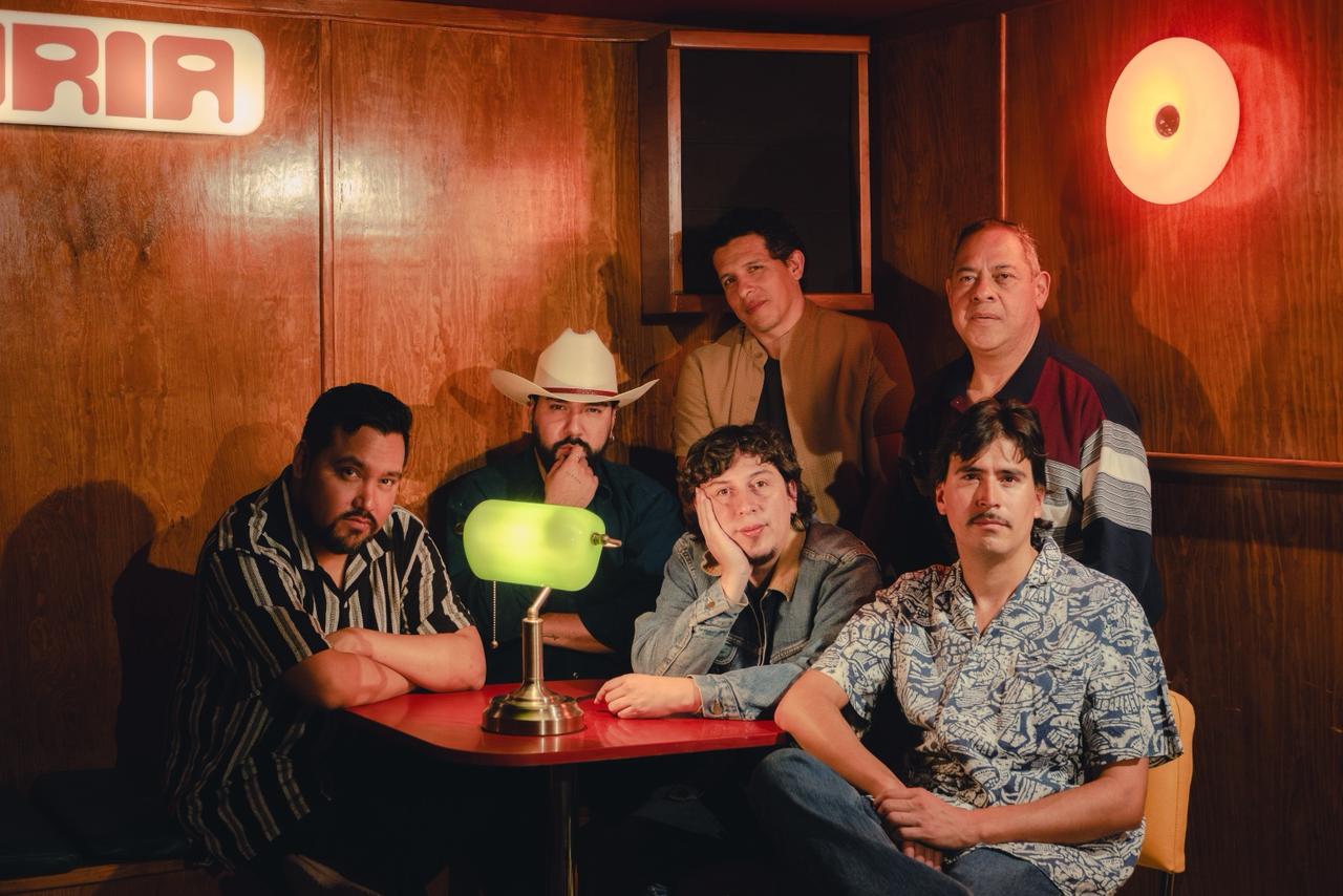 Un grupo de músicos posa alrededor de una mesa en un acogedor espacio de madera, iluminado por cálidas luces rojizas. La escena transmite camaradería y un ambiente íntimo, propio de un encuentro entre artistas.