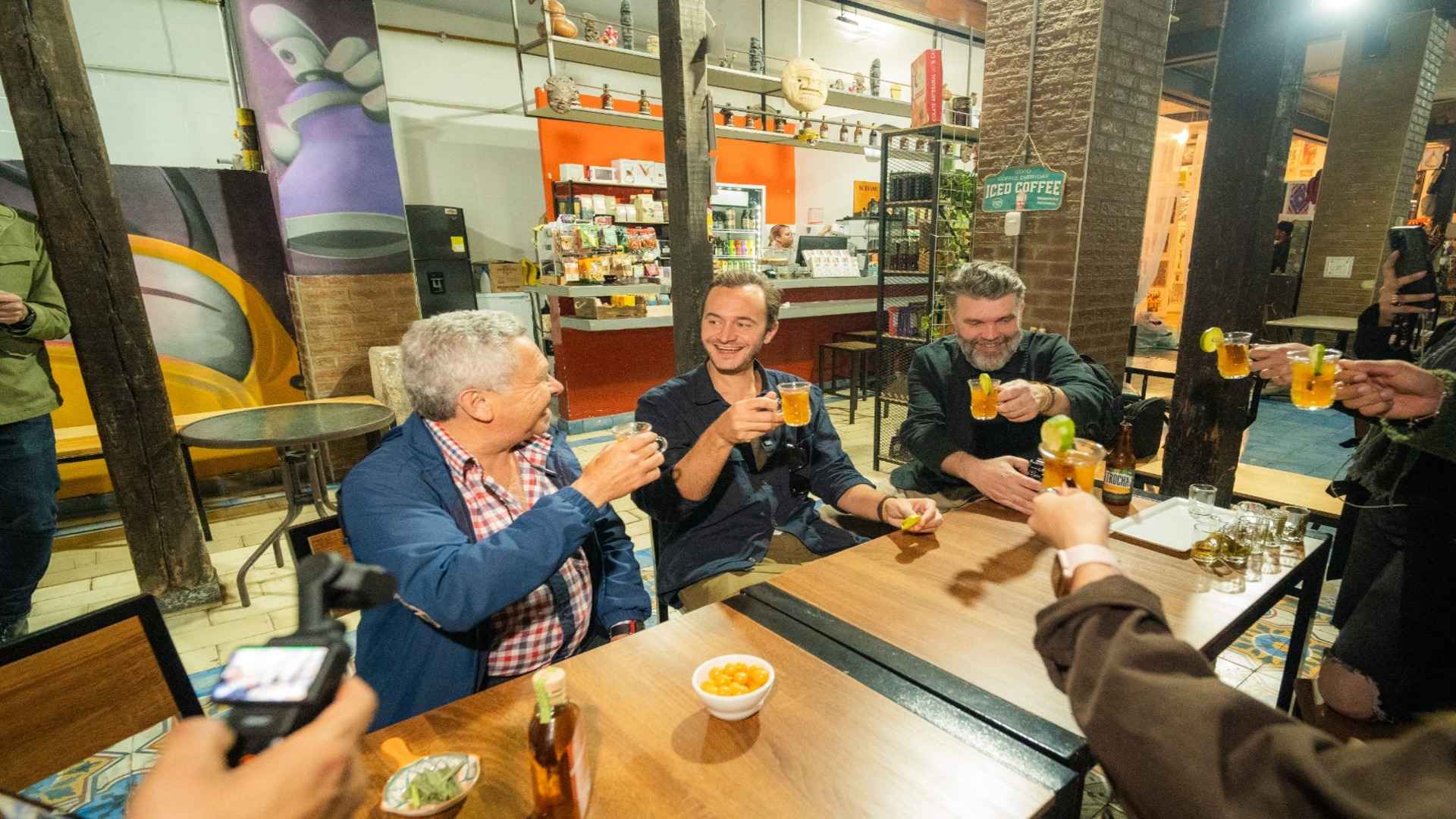 Grupo de personas alrededor de una mesa de madera brindan sonrientes con vasos de vidrio pequeños.