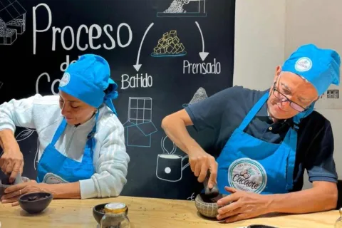 Un hombre y una mujer adultos con delantales y gorros azules muelen cacao en sendos morteros. Detás hay un aviso que señala el recorrido de la transformación del chocolate