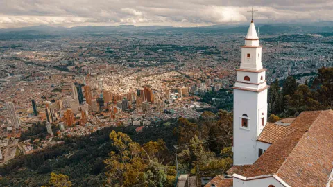 Cerro de Monserrate e primer plano de la torre de la iglesia y la montañan vista desde arriba con dron y al fondo la inmensa ciudad, sus edificios hasta el corredor de montañas del occidente. El cielo está tapado por nubes.