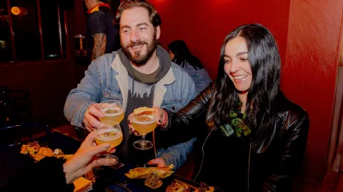 Un hombre y una mujer jóvenes brindan con copas con licor alrededor de una mesa con cómida típica colombiana. como empanadas, arepas y papas criollas.
