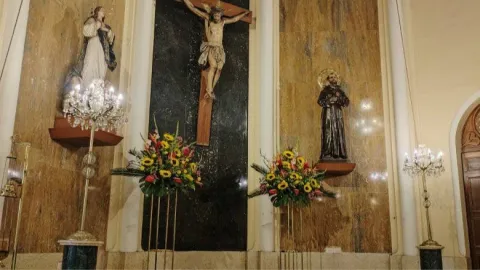 La Porciúncula Church, interior view of the nave with an image of the crucified Jesus, with the Virgin on one side and Saint Peter on the other. Below the figures on the wall, there are two floral arrangements and two multi-bulb lamps.