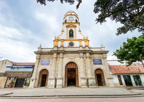  Iglesia Parroquial Santa Lucia - Chia con fachada frente con tres gigantes puertas en madera café  en contrapoicada con cielo azul y blanco. Un perro amarillo descansa al lado de una de las puertas.