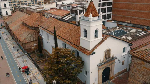 Iglesia de La Veracruz su fachada blanca y techo con tejas de barro, se observa abajo un jardín exterior con dos pinos, uno alto, un árbol frondoso y la carrera séptima con su ciclovía en la que hay algunas personas.  