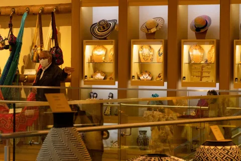 Gold Museum in the sales hall with wooden display cases showcasing handicrafts such as hats, vases, chains, hammocks, and bags. An elderly man wearing a tobacco hat points with his left hand.