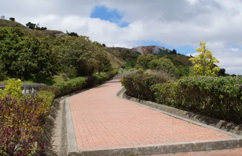 Parque Entrenubes con camino de ladrillo que conduce hacia arriba de la montaña rodeado por árboles pequeños y otra vegetación. El cielo está azul con algunas nubes.  