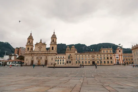 Plaza de Bolívar vista desde el occidente con algunas personas a lo lejos y de fondo la Catedral Primada y el Sagrario. Al fondo los cerros orientales y el cielo blanco con dos palomas en vuelo.