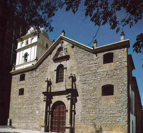 Church of St. Augustine, front view with a colonial gray stone facade, wooden door, windows with iron grilles, and a yellow bell tower ending in a dome. Tree branches decorate the view under a blue sky.