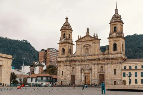 Catedral Primada de Colombia, plano general fachada vista desde el sur occidente con personas en la Plaza de Bolívar, la Casa de la Independencia y los Cerros Orientales de fondo. El cielo es blanco.