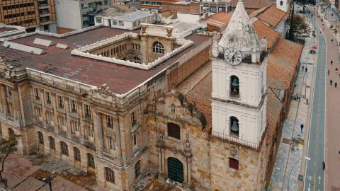 Iglesia San Francisco colonial en primer plano de fachada con blanca torre del reloj y techo, al lado se observa la cicloruta de la carrera séptima con algunas personas caminando.