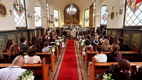 Iglesia Santa Mónica al interior con celebración eucarística, sillas repletas de personas y al fondo el sacerdote da la comunión a un niño. En las paredes resaltan los vitrales religiosos.