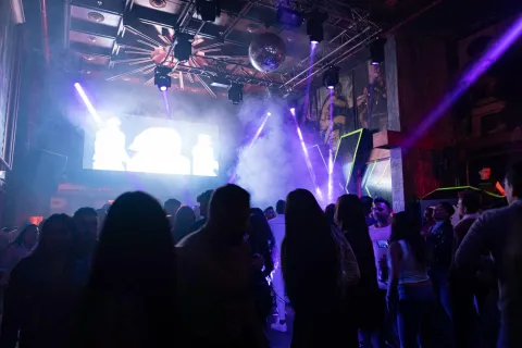 Theatron interior en sala de eventos oscura con iluminación de refletores, una pantalla gigante y sombras de varias personas que bailan y conversan