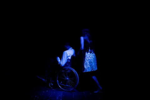  Barraca Teatro con actor en silla de ruedas en escena con luz azul. Una mujer lo mueve en la silla.