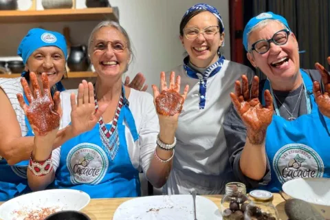 Cuatro mujeres, tres adultas y una joven con delantales y gorros azules sonríen y muestran sus manos untadas de cacao.