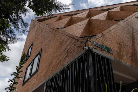 Chapinero con fachada de edificio con forma geométrica y en ladrillo con dos ventanas cuadradas y rejas metálicas color negro. A la izquierda hay unas ramas de árbol.
