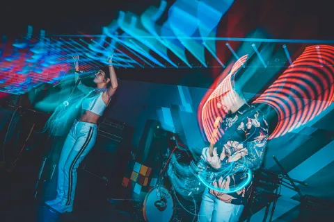 Disco Jaguar al interior en escenario con dos musicos mujer y hombre jóvenes cantando e iluminados con luces psicodélicas azules y rojas.En la decoraciuón hay un cubo de Rubik gigante.