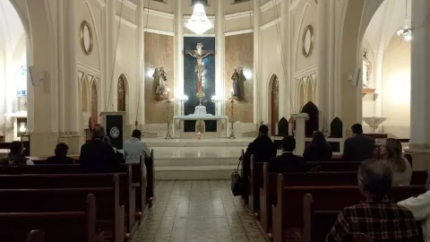  La Porciúncula Church, interior with people praying, sitting, and kneeling in front of the altar. The walls decorated with circles and ovals stand out, as well as a large lamp hanging directly above the figure of Christ.