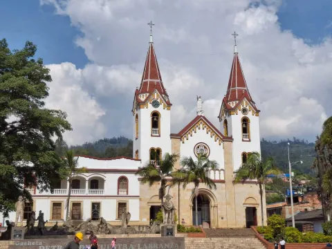Iglesia Parroquial San Miguel Arcángel - Gachetá fachada de frente en parque rodeada de árboles  y obreros que caminan al frente. Al fondo se observan montañas y un cielo azul con nubes blancas.