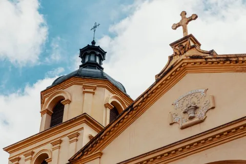  Iglesia de la Candelaria con la parte alta de su fachada en primer plano, se resalta la punta del frente coronada por una cruz y una torre con su cúpula negra y una cruz metálica encima. 