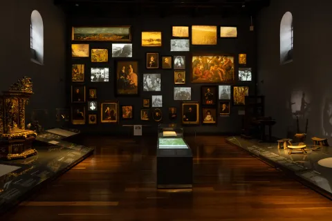 National Museum of Colombia, interior exhibition hall with a back wall covered with photographs and paintings. Display cases feature items such as historically valuable chairs. The room is lit with artificial light, complemented by natural light coming through two small windows high on the left and right walls.