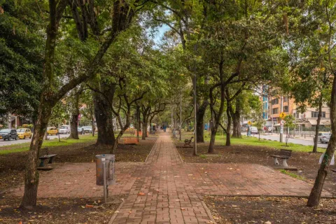 Park Way dentro del corredor arborizado con un camino de ladrillos y una silla de madera y concreto a lado y lado y una caneca metálica colgada de un árbol. A la ixquierda se ve una calle concurrida por vehículos particulares y taxis.