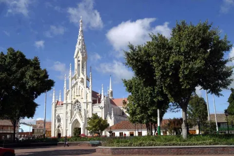 Parroquia Basílica Menor del Santo Cristo - Ubaté neogótica en parque rodeada de árboles y cielo azul con una nubecita blanca. Se en dos personas caminando a lo lejos.