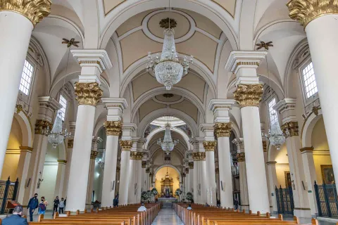 Catedral Primada de Colombia, interior vista desde la puerta hacia el altar de fondo con muchas filas de sillas con pocas personas sentadas. Destacan 7 grandes columnas blancas a cada lado, los ventanales y 2 gigantes lámparas que cuelgan del techo. 