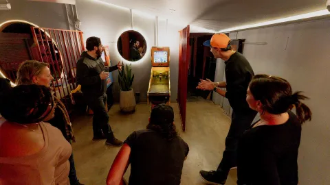 Grupo de hombres y mujeres en un salón jugando Bolirana. Todos observan con emoción la maquina de juego.