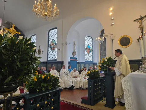  Iglesia Santa Mónica al interior primer plano del sacerdote en el atrio que habla al microfono para un grupo de sacerdotes. Lo rodean cuatro arreglos florales, un Cristo a espaldas, una Virgen María al frente y dos lámparas que cuelgan del techo.