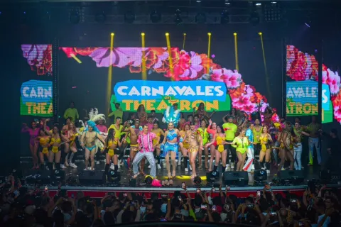 Theatron vista clara de escenario con decenas de artistas disfrazados con trajes de colores y un título que dice Carnavales en Theatron. Al frente de la tarima se alcanza a ver a un grupo de personas que levantan la mano.