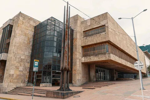Luis Ángel Arango Library, modern section with a façade of brown tiles, a large glass door, and a metal monument. There is a SITP bus stop.